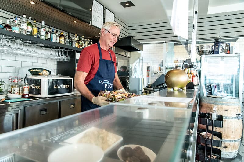 Aelterer Mann mit grauen Haaren und Brille steht hinter einer Theke in einer Bar, traegt ein rotes Shirt und eine blaue Schuerze und stellt einen Teller mit Essen ab; hinter ihm stehen Flaschen und Glaeser.