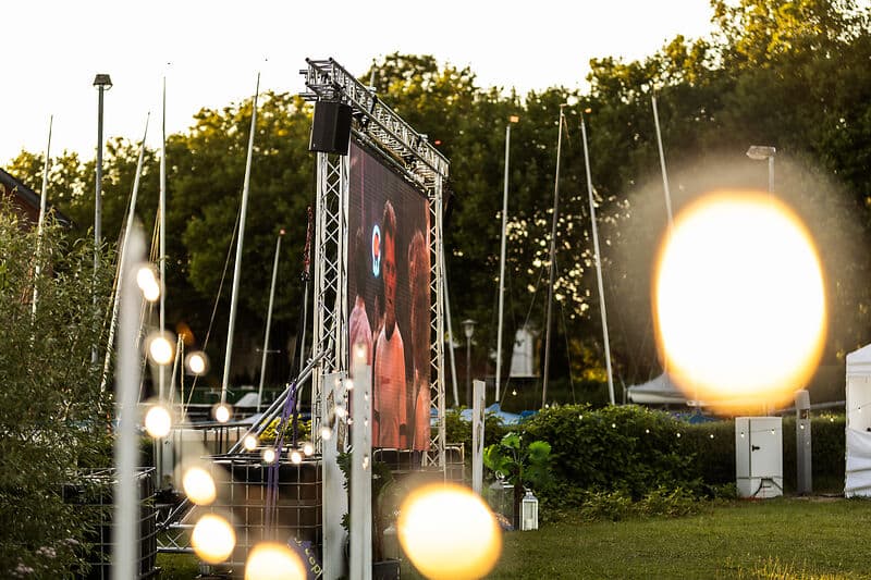 Große LED-Leinwand auf einem Metallgerüst im Freien; darauf sind zwei Menschen zu sehen. Im Vordergrund hängen unscharfe, leuchtende Lichter.