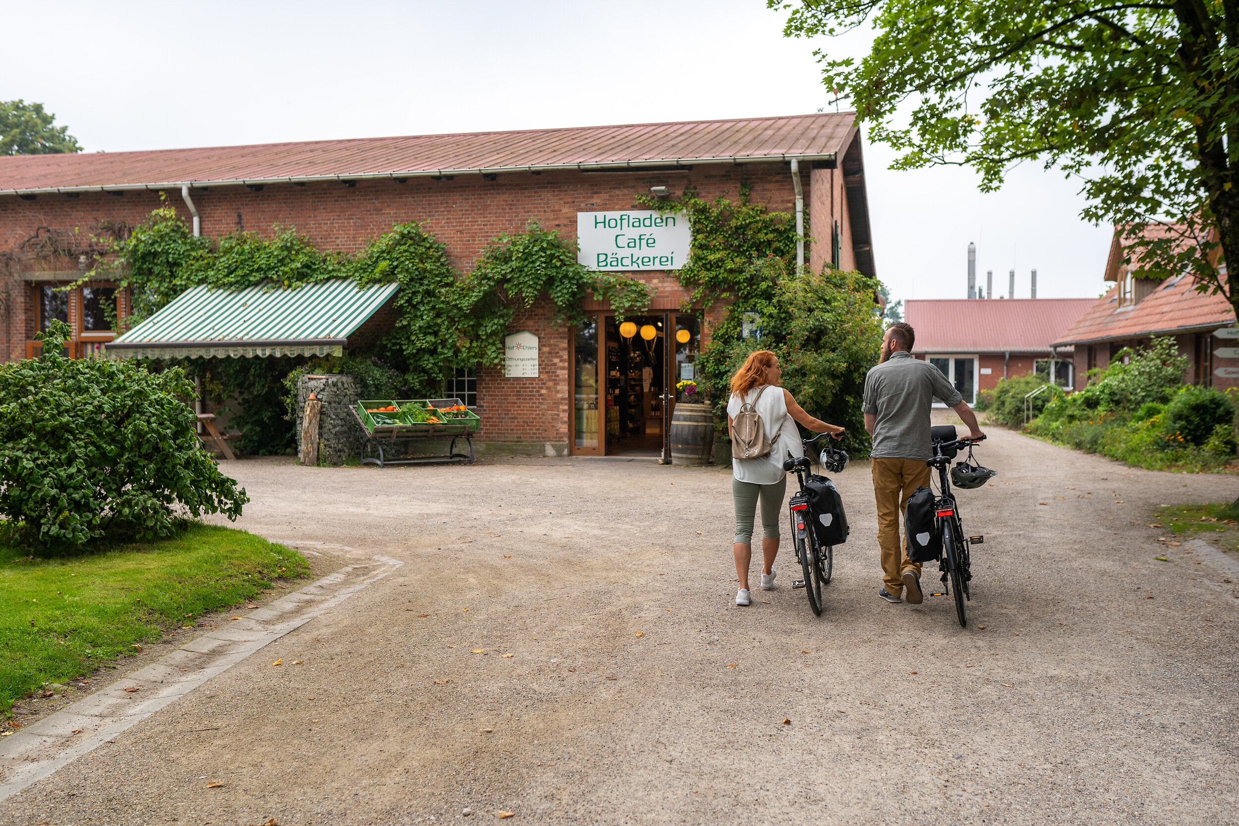 Zwei Radfahrer schieben ihre Räder auf den Hof vor einem Hofladen mit Café und Bäckerei