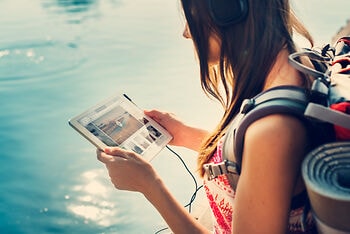 Frau mit Rucksack und großen Kopfhörern steht am Wasser und hält ein Tablet in beiden Händen.