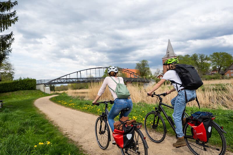 Zwei Radreisende mit Helmen fahren auf einem Feldweg Richtung Brücke; beide Fahrräder haben Gepäcktaschen, im Hintergrund steht ein Kirchturm unter wolkigem Himmel.