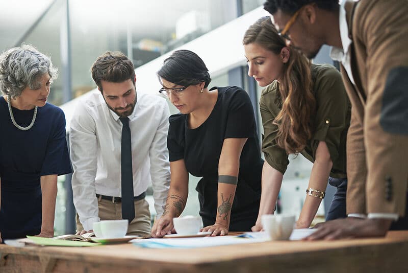 Mehrere Personen stehen in einem Büro um einen Tisch und beugen sich über große Pläne und Papiere, als würden sie gemeinsam etwas besprechen.