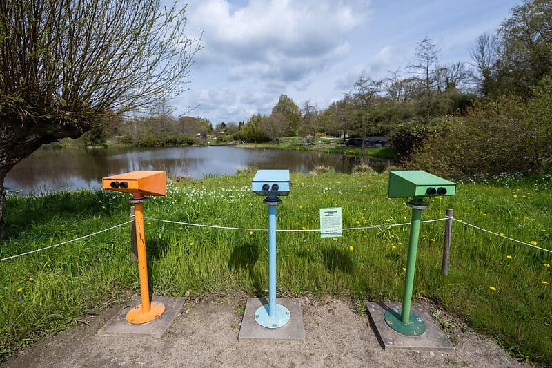 Drei fest montierte Fernrohre in Orange, Blau und Grün stehen auf einer Wiese am Seeufer; dahinter ein ruhiger See und Bäume unter wolkigem Himmel.