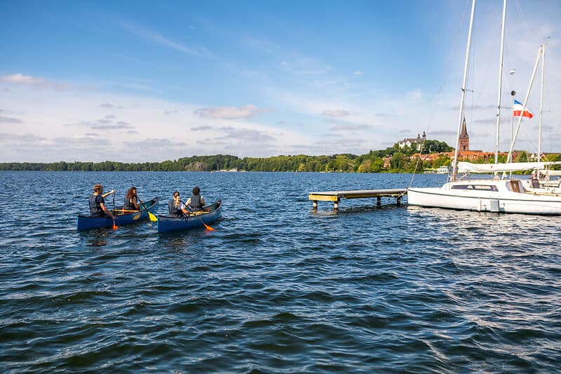 Zwei Kanus mit insgesamt fünf Personen paddeln auf einem See; rechts liegen ein Steg und ein weißes Segelboot, im Hintergrund ein Ort mit Kirchturm.