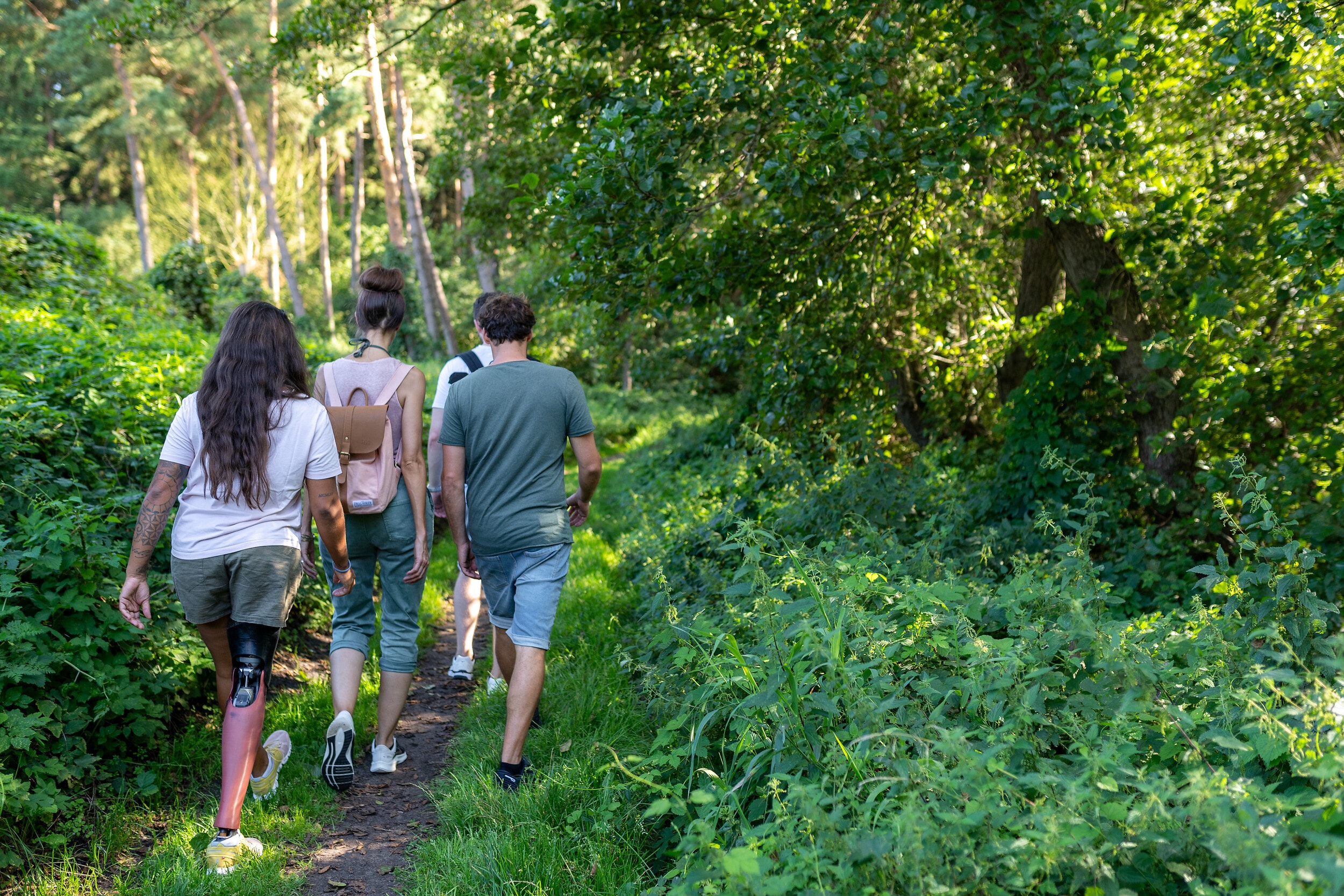 Vier Personen, darunter eine Person mit Beinprotese, spazieren über Trampelpfad durch den Wald