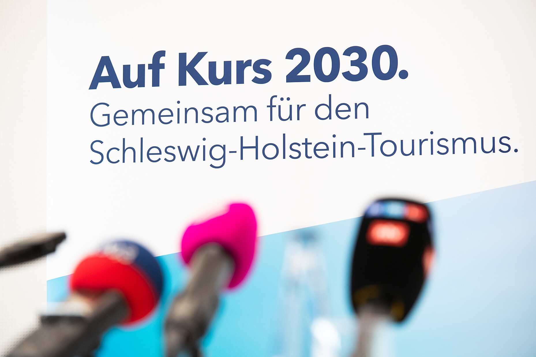 Schriftzug "Auf Kurs 2030 - Gemeinsam für den Schleswig-Holstein-Tourismus" vor Mikrofonen bei einer Pressekonferenz