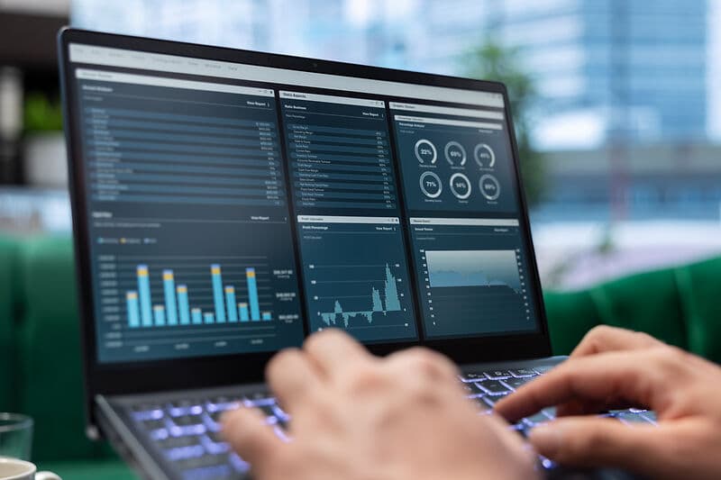 Hände tippen auf einer Laptop-Tastatur; auf dem Bildschirm sind mehrere Diagramme und Auswertungen in einem dunklen Dashboard zu sehen.